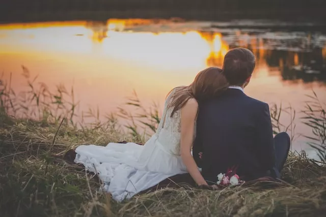 As the golden hour bathes the lake in a warm glow, the honeymoon couple sits together, the woman leaning on the man's shoulder. It's a beautiful moment to cherish.